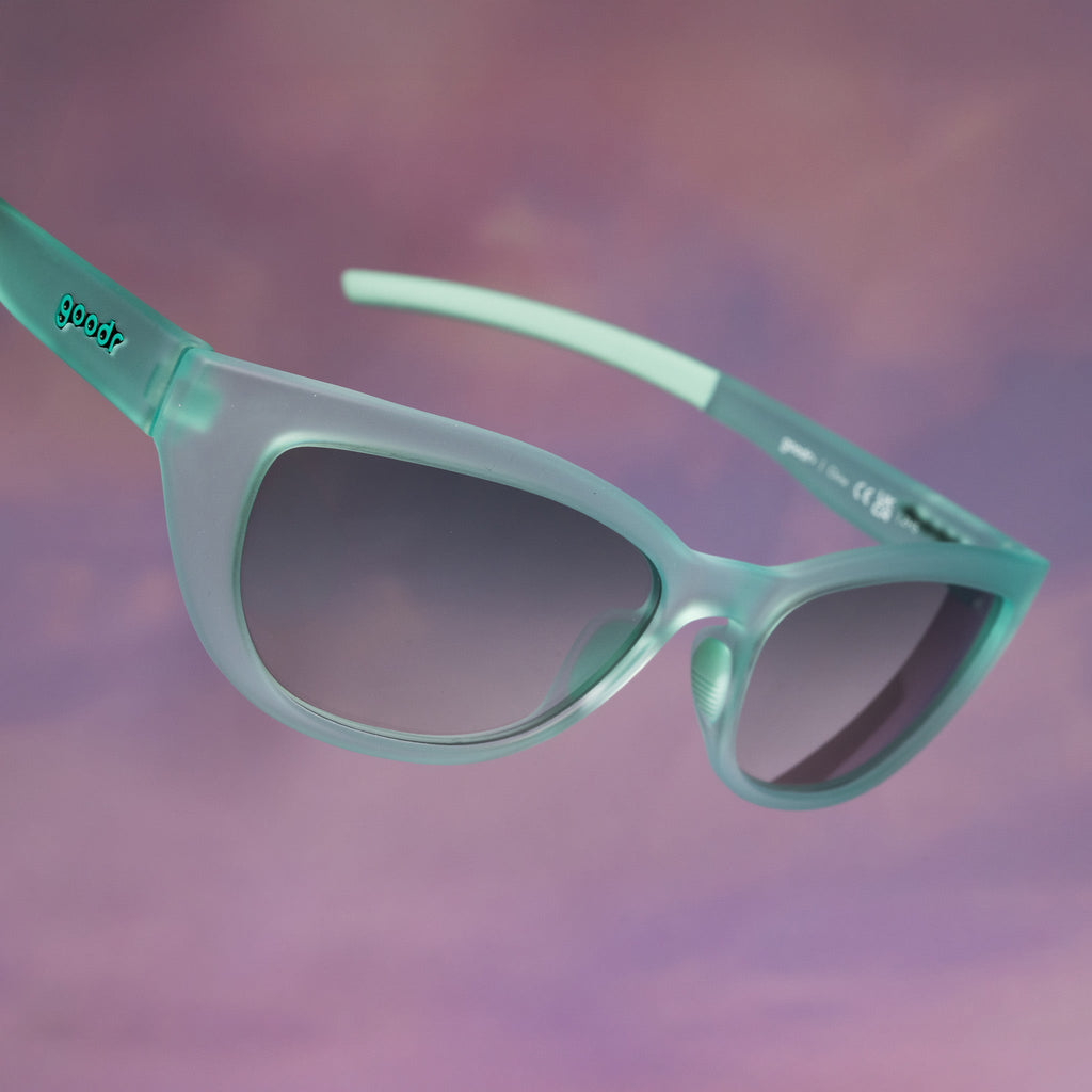 Three-quarter angle view of translucent pale green Cheetah G sunglasses floating against a pinky peach ombre backdrop.