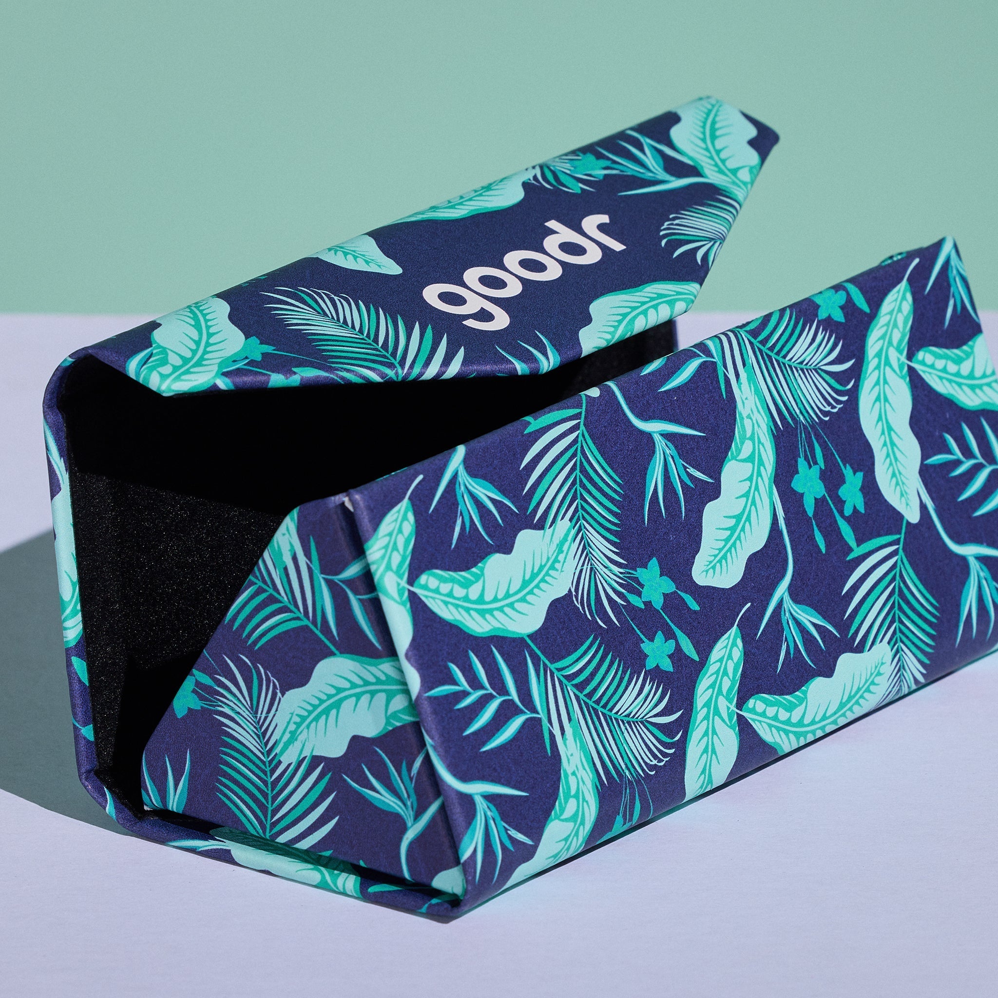 Close-up view of the Enter the Fold navy and teal tropical-print sunglasses case showing it expanded to hold a pair of sunglasses.
