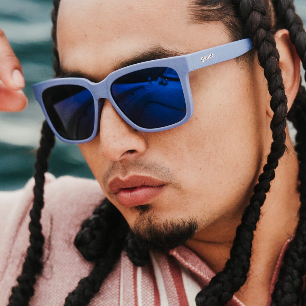 Three-quarter angle view of a composed young man with braids wearing dark blue sunglasses with dark blue lenses.