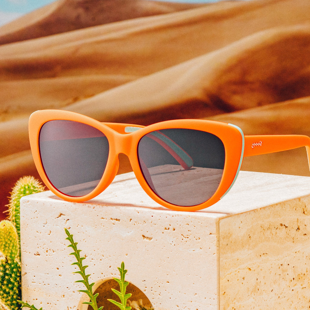 Three-quarter angle view of red cat-eye sunglasses with black non-reflective lenses on top of a clay block against a desert backdrop.