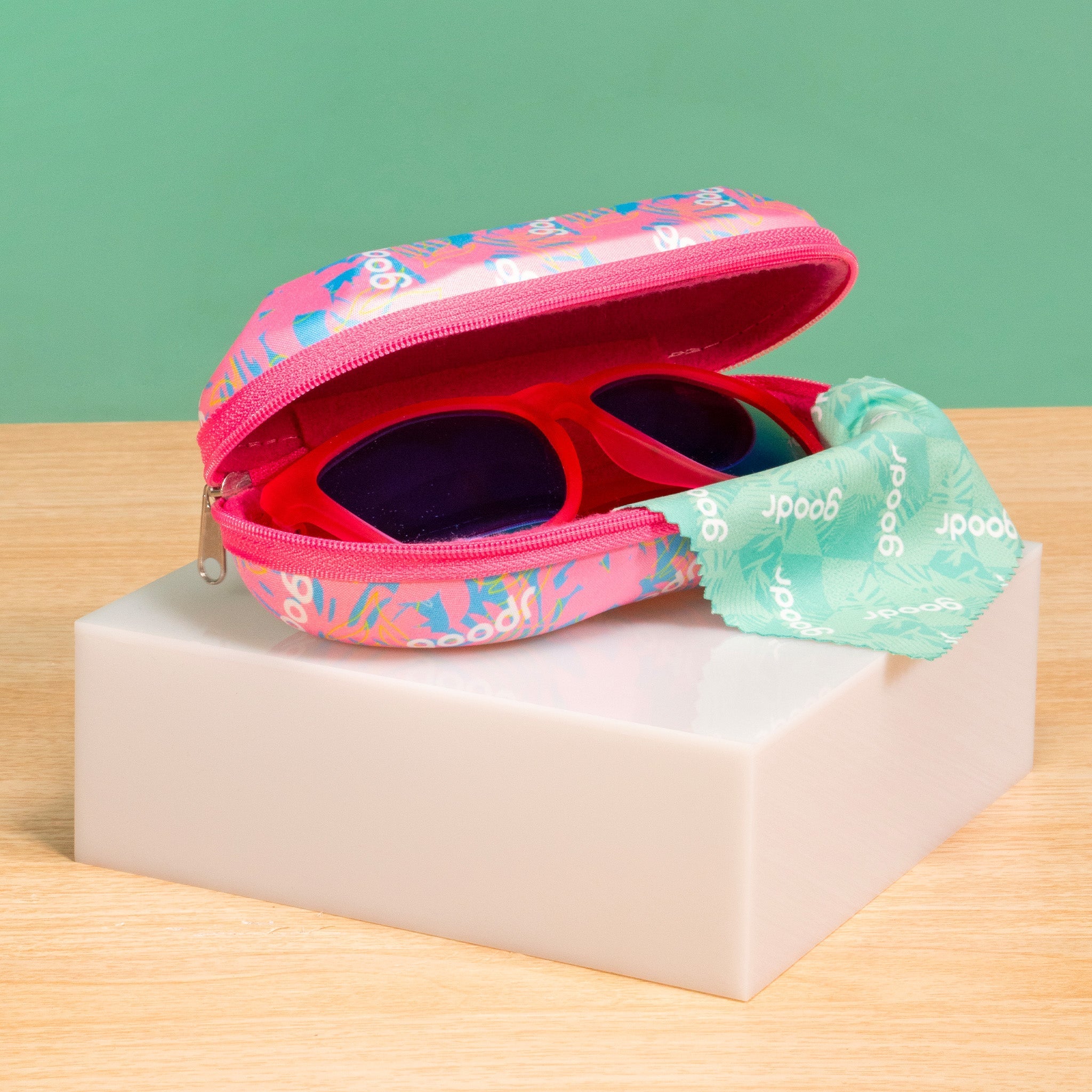A pair of pink goodr sunglasses sitting in a pink, teal, and white branded goodr case, on top of a small white platform on a table.