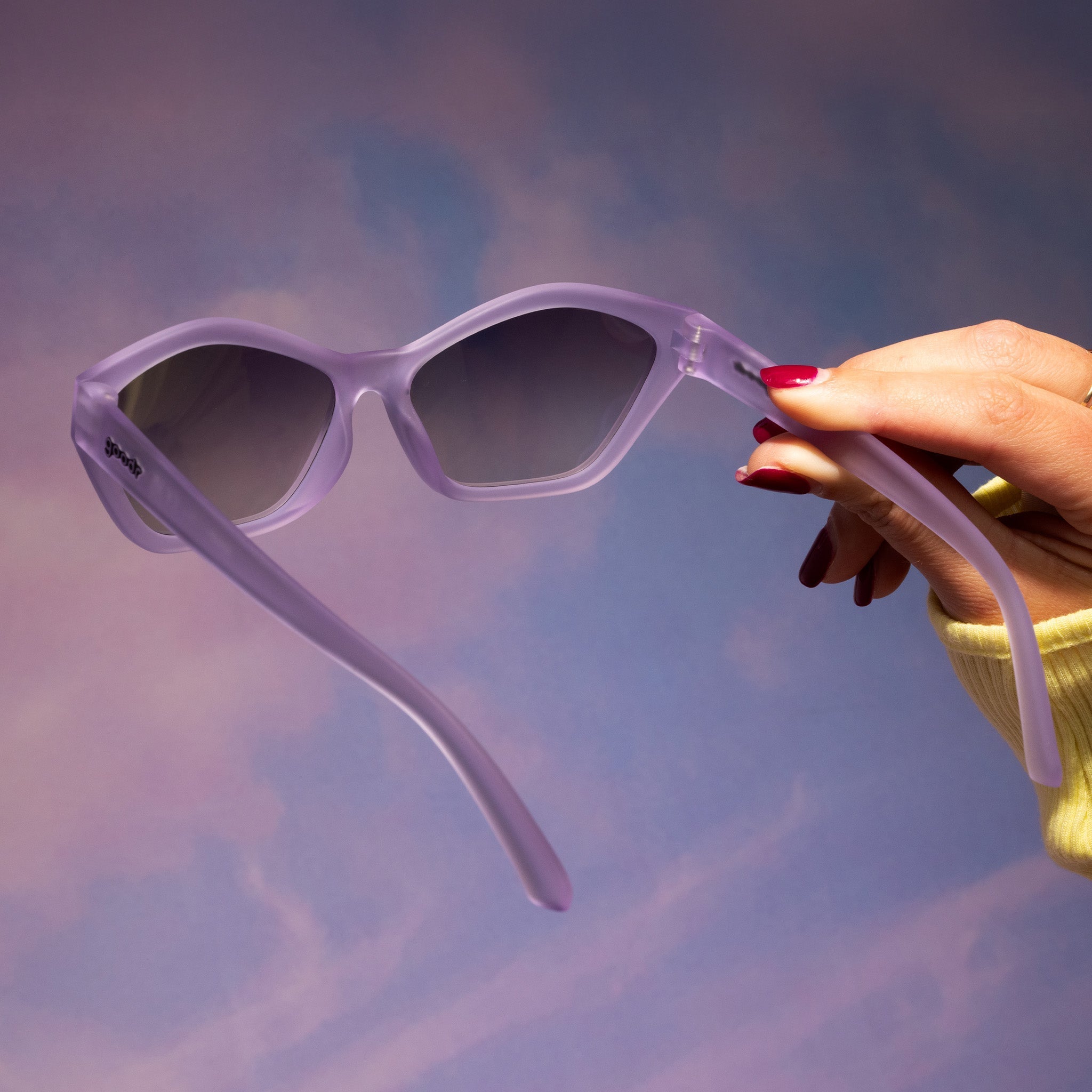 A hand with red fingernails holds up a pair of translucent pale purple Deco G sunglasses against a purplish pink backdrop.