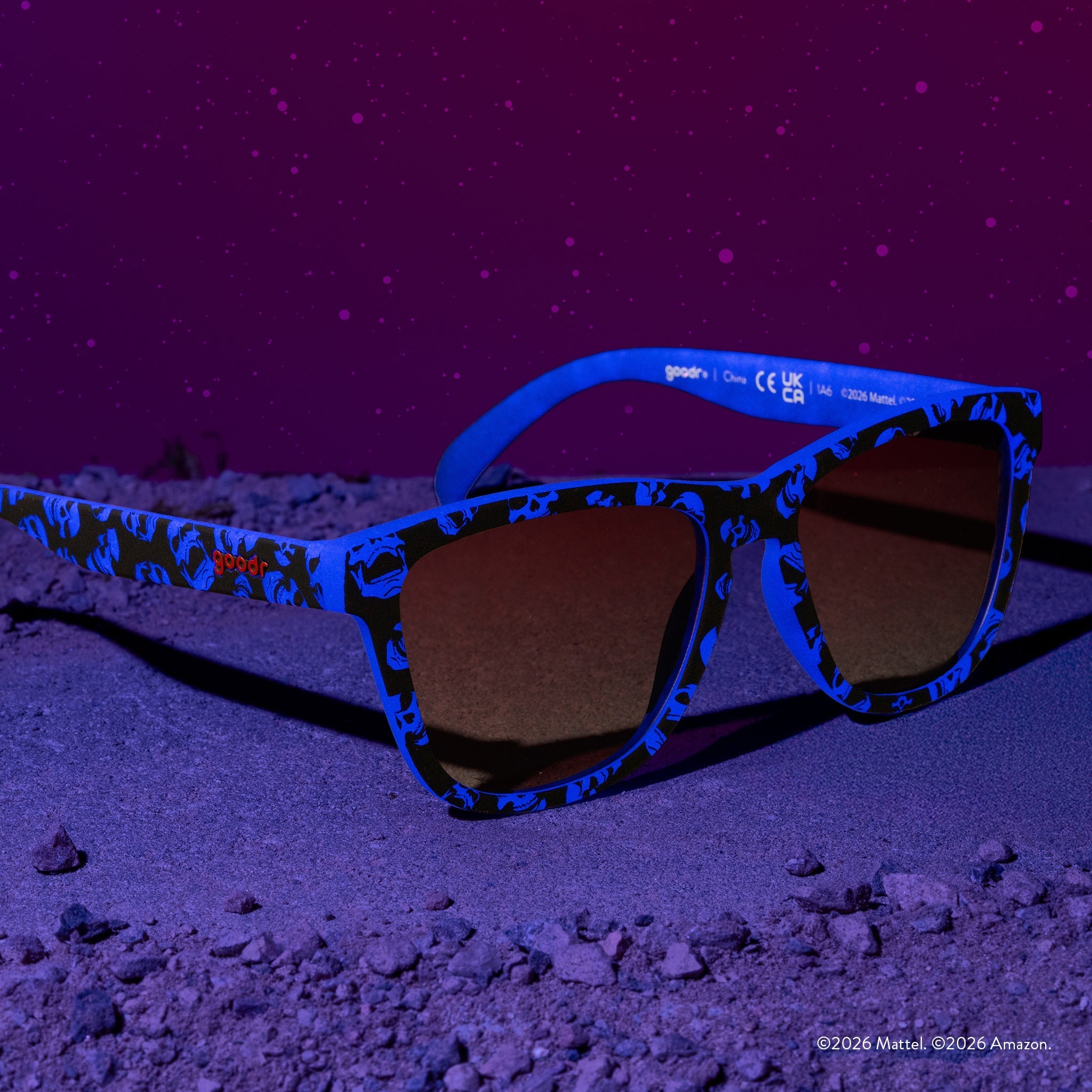Three-quarter angle view of blue and black sunglasses featuring a Skeletor pattern and rose gradient lenses, set against a low-lit purple background.