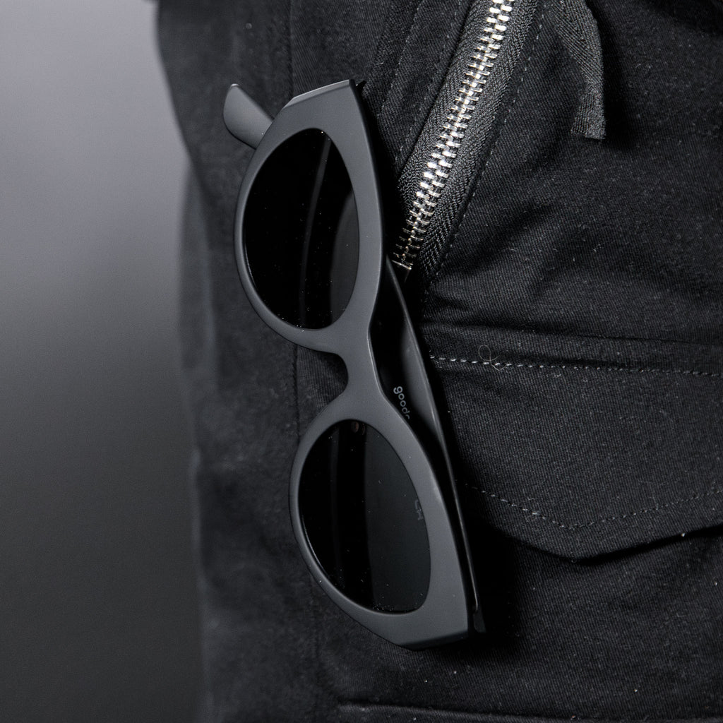 Close-up of translucent black, retro-inspired angled cat-eye sunglasses with non-reflective lenses hanging from a black zippered pant pocket.