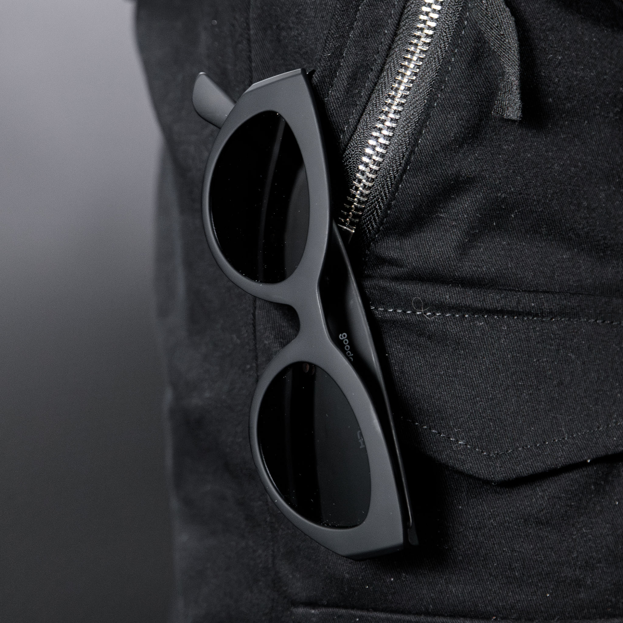 Close-up of translucent black, retro-inspired angled cat-eye sunglasses with non-reflective lenses hanging from a black zippered pant pocket.