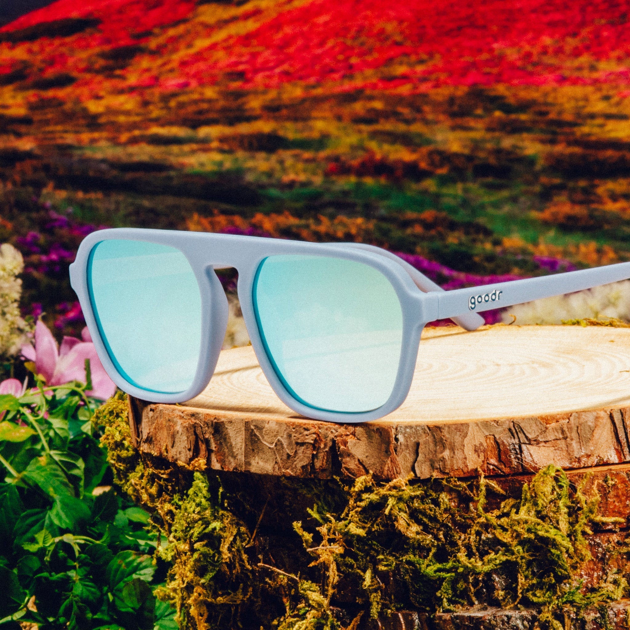 Three-quarter angle view of light blue retro-inspired aviator sunglasses with blue reflective lenses on a tree trunk against a meadow backdrop.