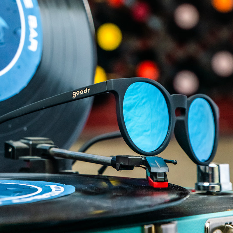 A pair of round sunglasses with black frames and blue lenses, sitting on a record player.
