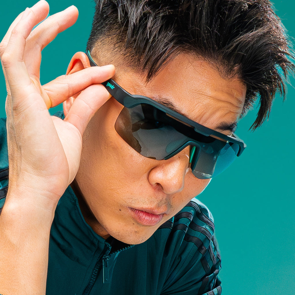 A man with dark hair playfully grasps the temple of his dark teal Bolt Gs with one hand as if he's about to tip them down to reveal his eyes.
