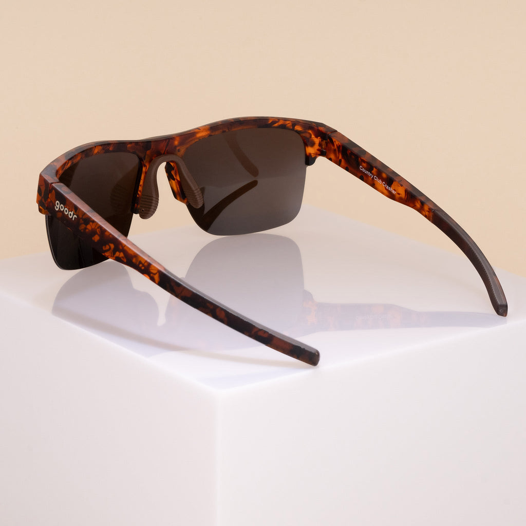 Three-quarter back angle view of half-rim brown tortoiseshell sunglasses with brown non-reflective lenses. The arms are extended, showcasing the goodr logo on the outer left arm, an adjustable brown silicone nose piece, and brown silicone grips on the inner arms.