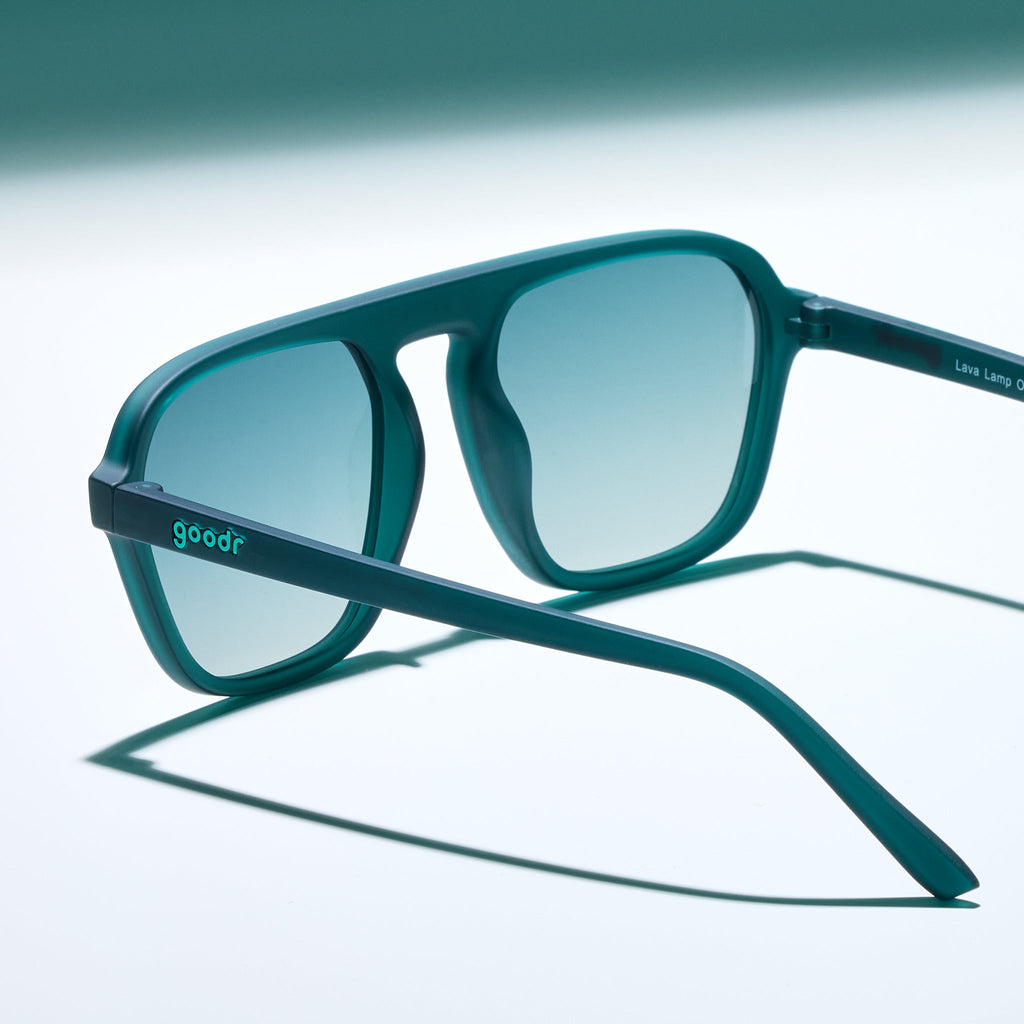 Three-quarter angle view of a pair of translucent teal Retro Gs with green gradient lenses sitting on a white surface, photographed from behind.