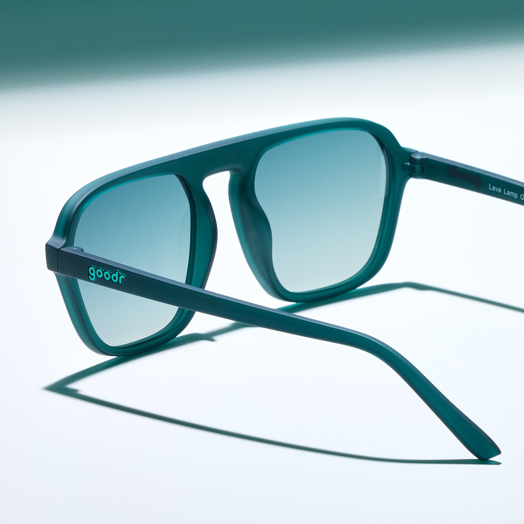 Three-quarter angle view of a pair of translucent teal Retro Gs with green gradient lenses sitting on a white surface, photographed from behind.