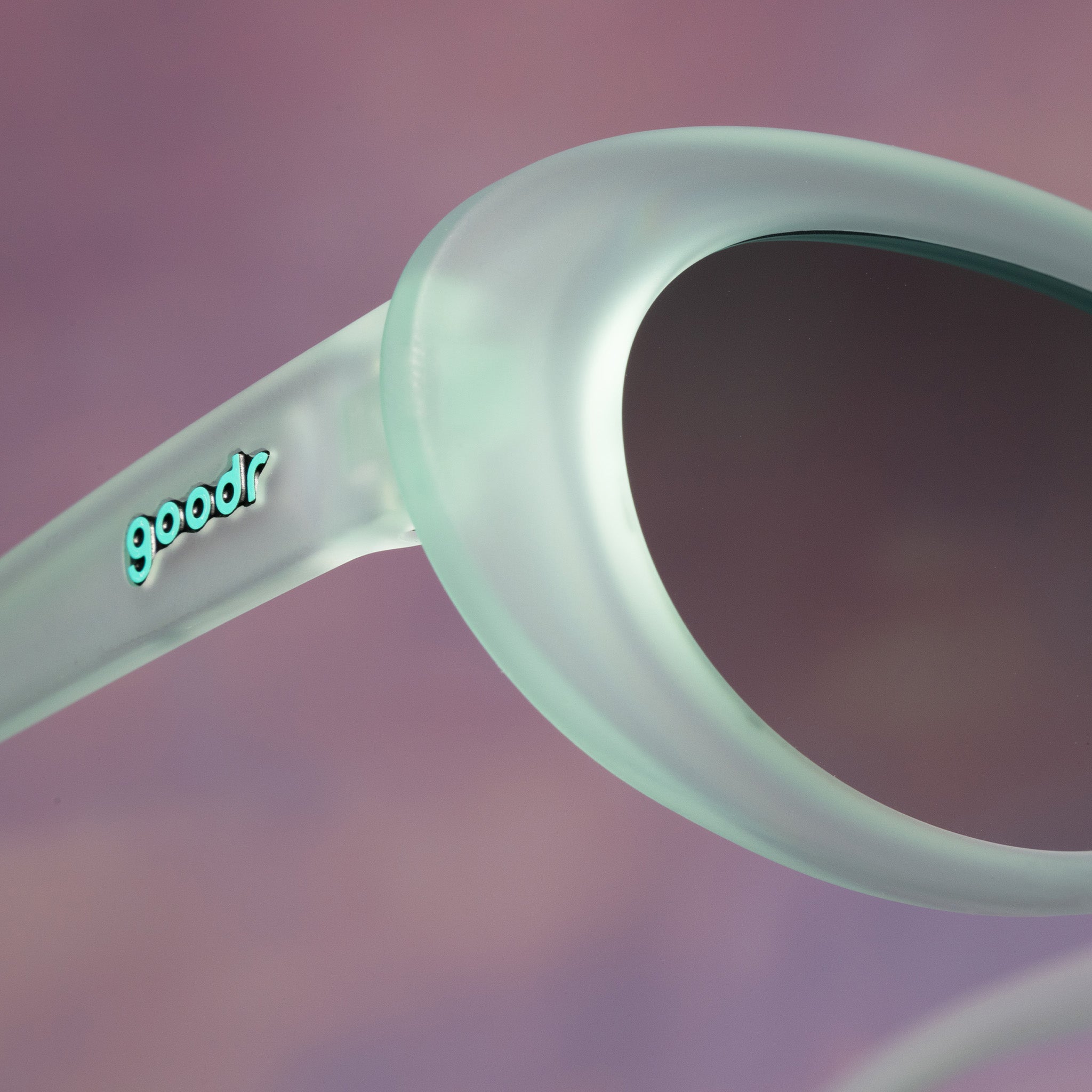 Close-up of the corner temple of a pair of pale green oval Beat G sunglasses against a pale purple-pink backdrop.