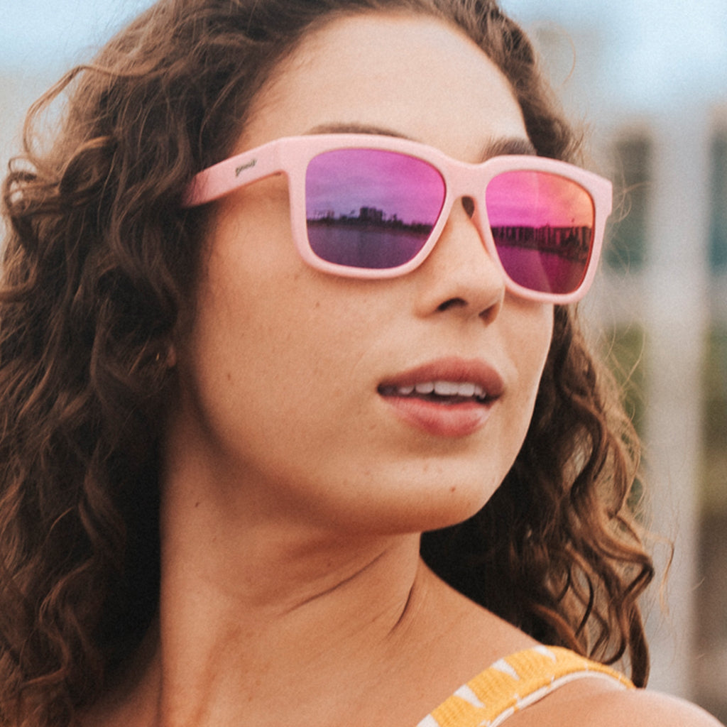 Three-quarter angle view of an amazed young woman with dark hair wearing pink sunglasses with pink-purple lenses.