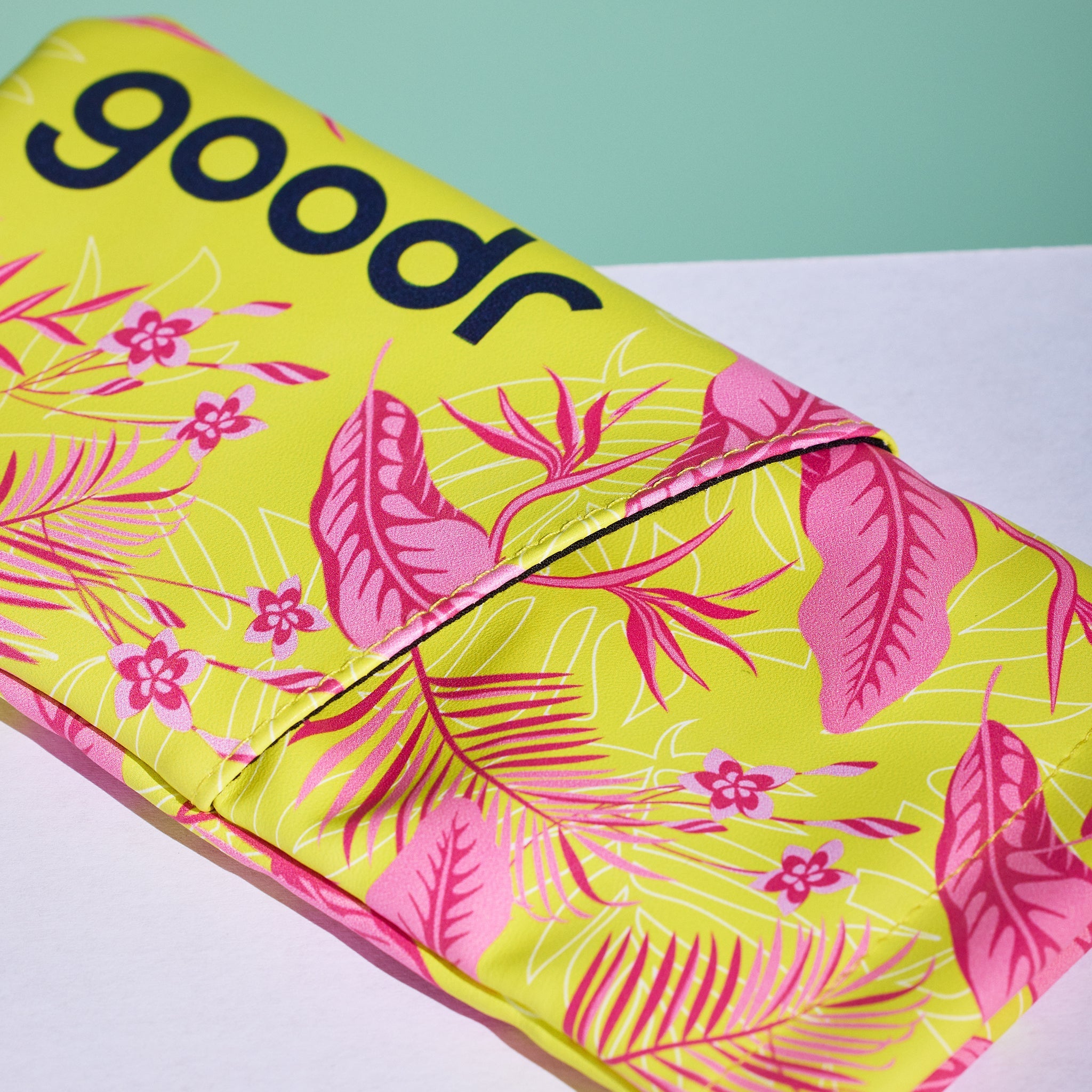 A close-up view of a vibrant pink and yellow tropical print fabric sunglasses case displayed on a white surface with a sage green backdrop.