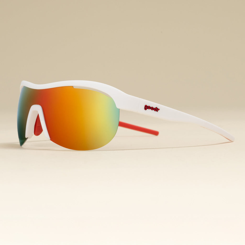 Left three-quarter angle view of white wrap shield sunglasses with red accents and fire-red reflective lenses on a tabletop.