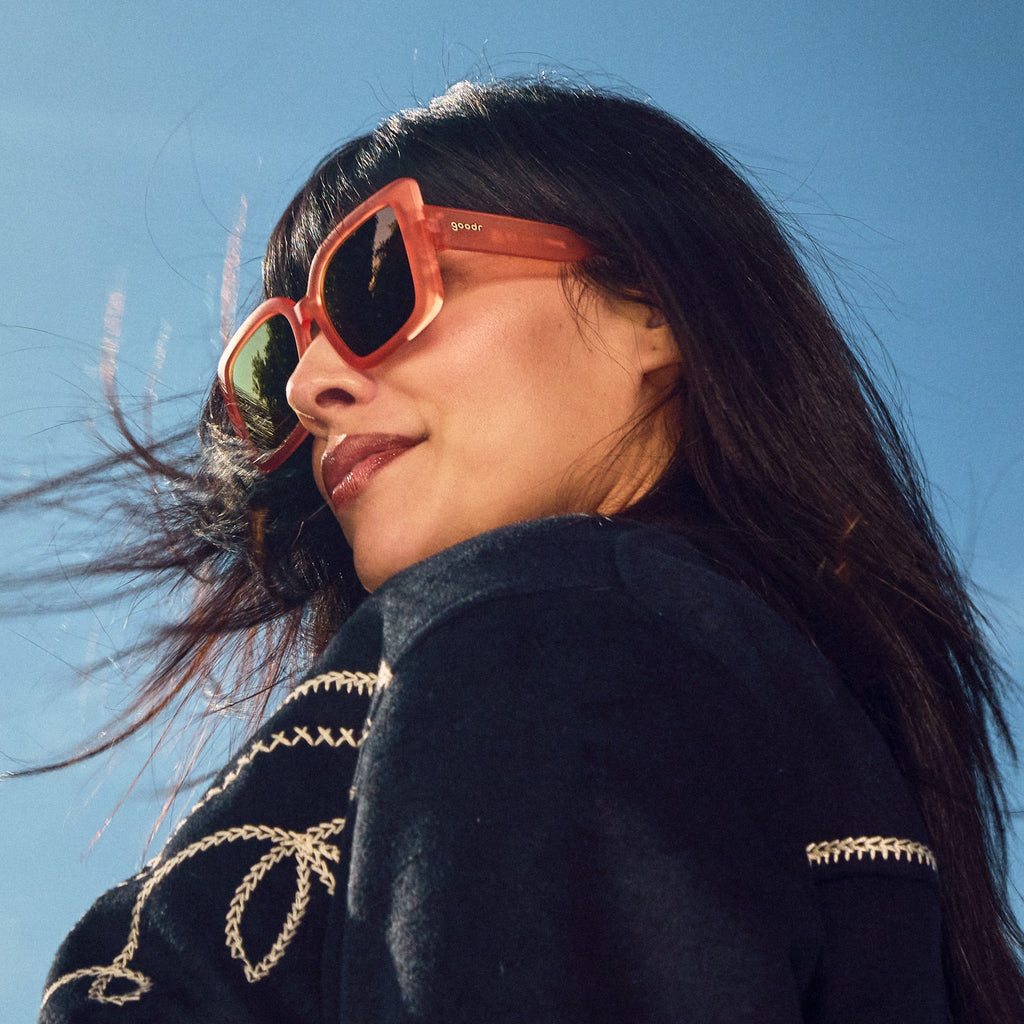 Low-angle view of a woman posing, wearing oversized square sunglasses in coral pink with champagne gold reflective lenses.