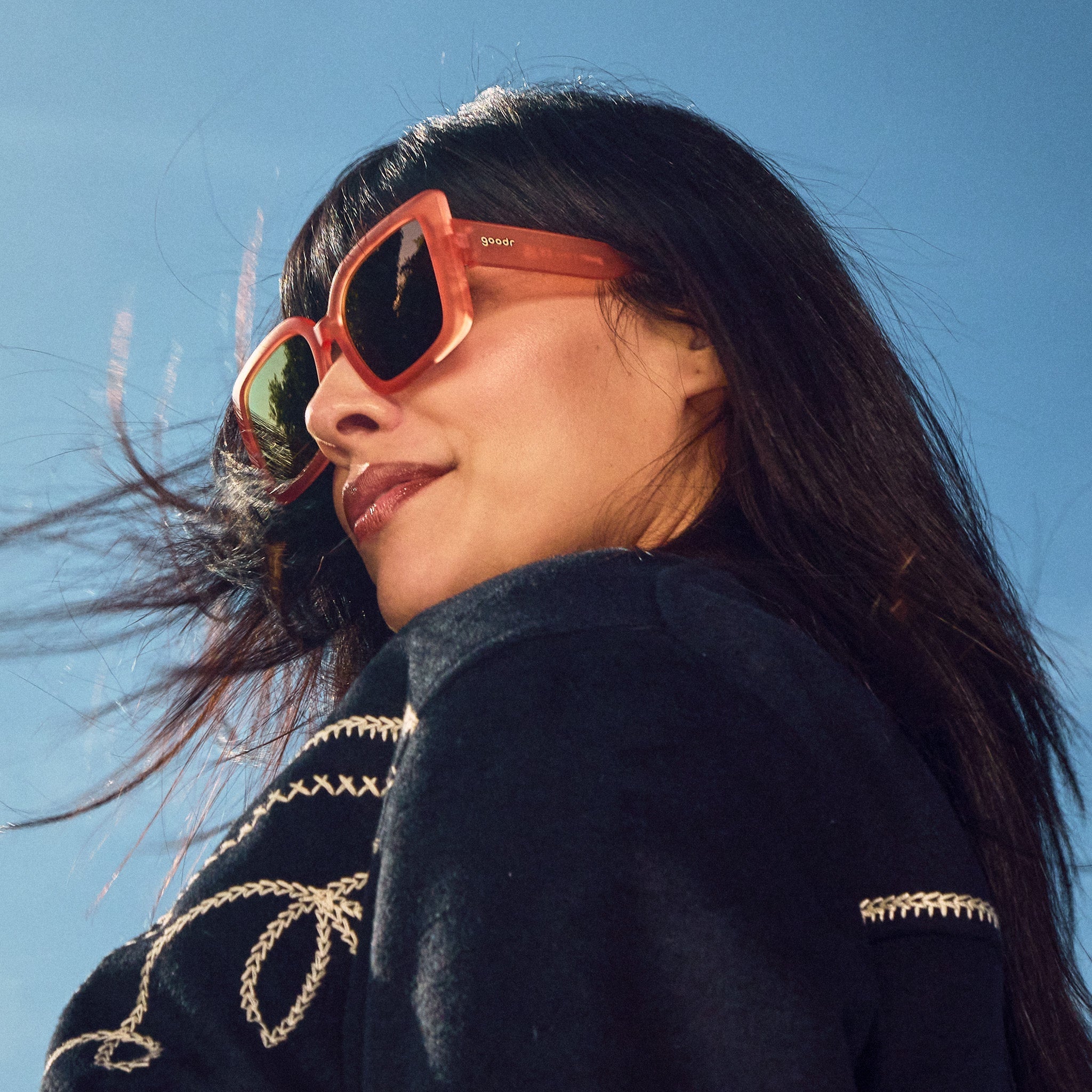 Low-angle view of a woman posing, wearing oversized square sunglasses in coral pink with champagne gold reflective lenses.