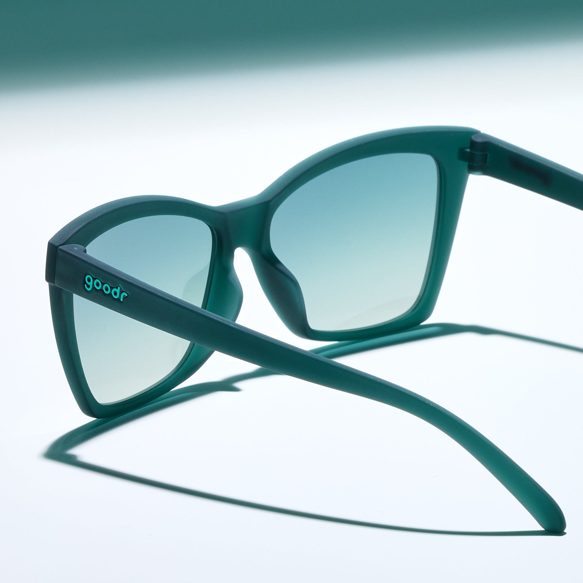 Three-quarter angle view of a pair of translucent teal Pop Gs with green gradient lenses sitting on a white surface, photographed from behind.