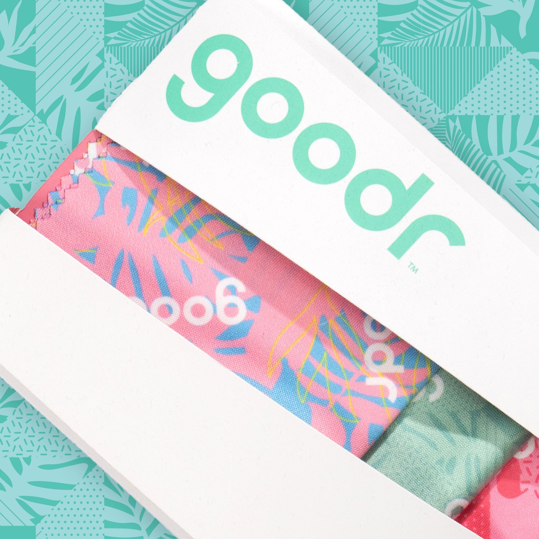 Close-up of a white box housing a pack of three tropical microfiber lens cloths in shades of pink and teal.