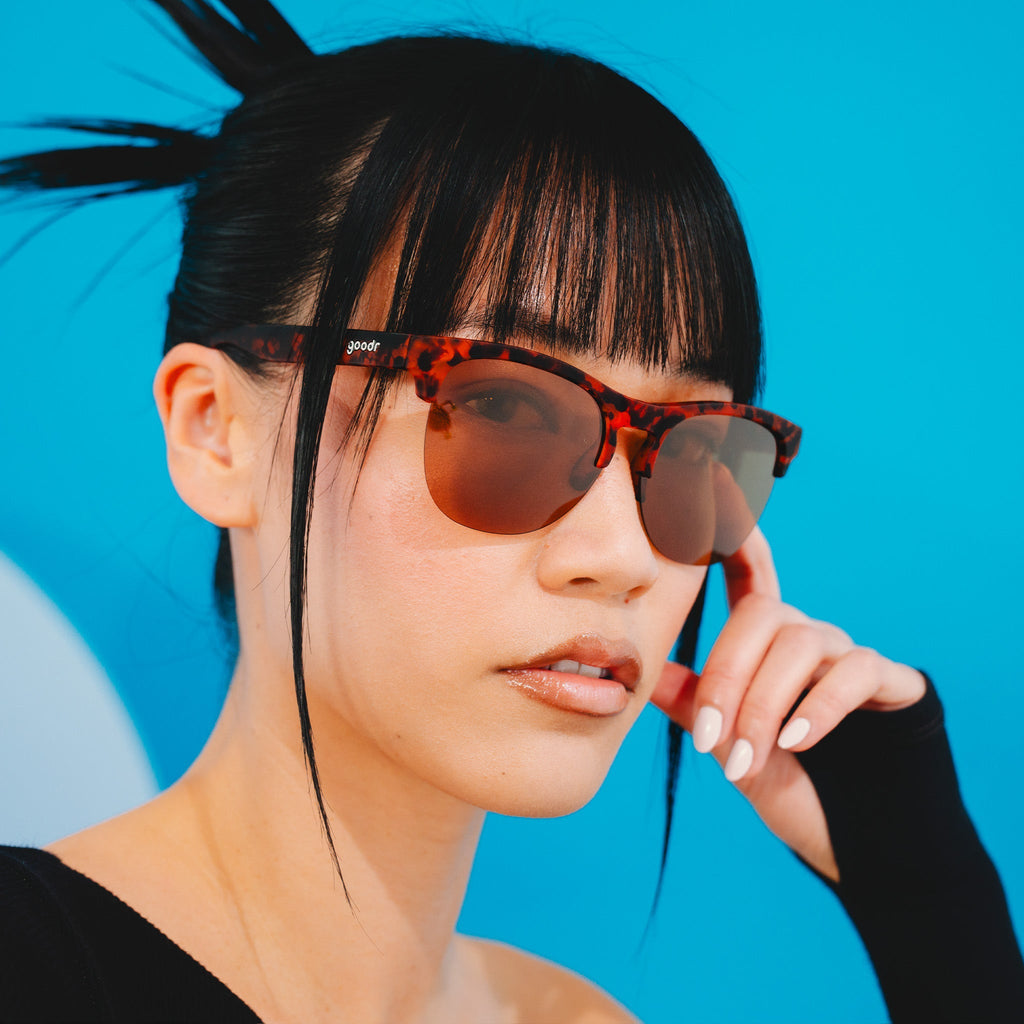 A stylish woman wearing brown tortoiseshell half-rim sunglasses with brown non-reflective lenses poses for the camera.