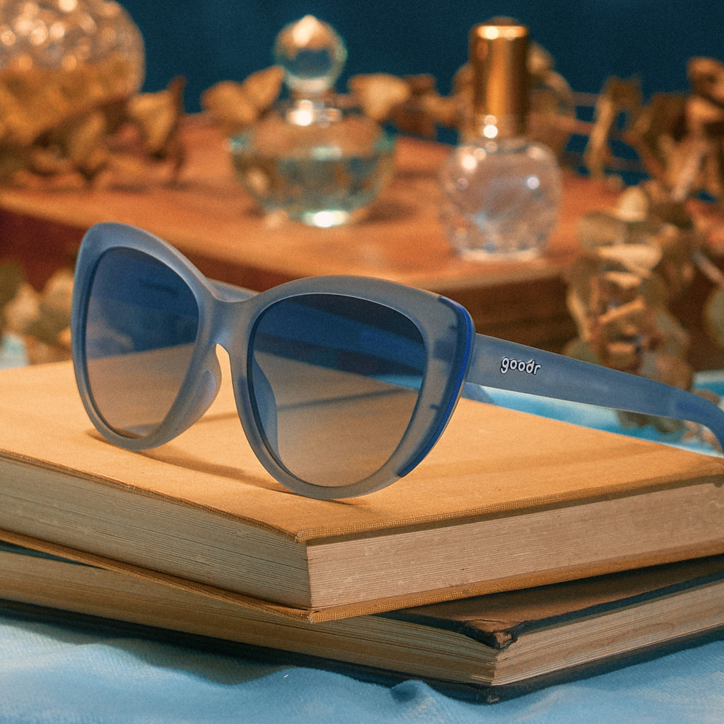A pair of dusty blue translucent Glam Gs sits atop some vintage books, with dried flowers and perfume in the background.