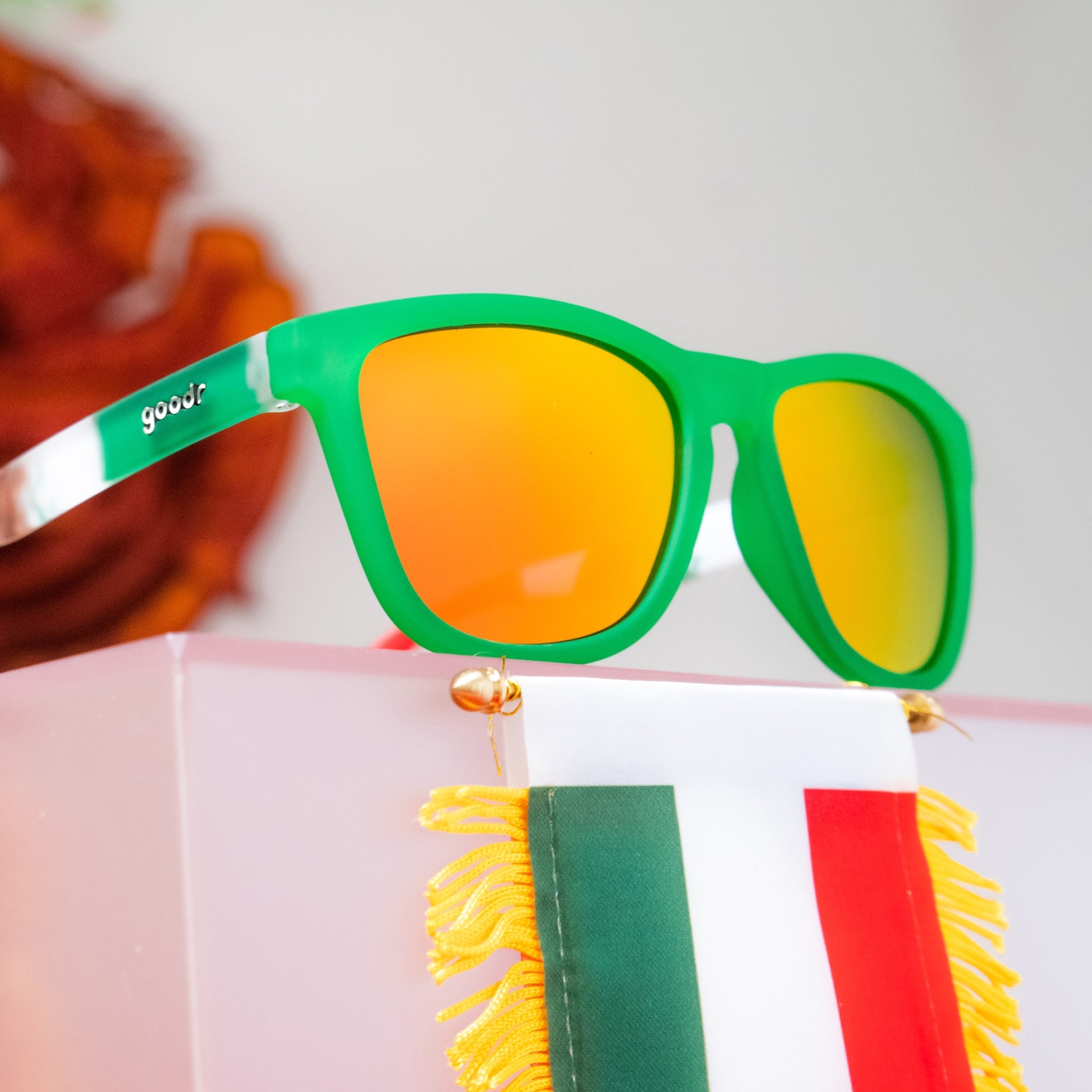 Angled view of green shades with a Mexican flag pattern+burnt orange reflective lenses on a podium with a Mexico flag.