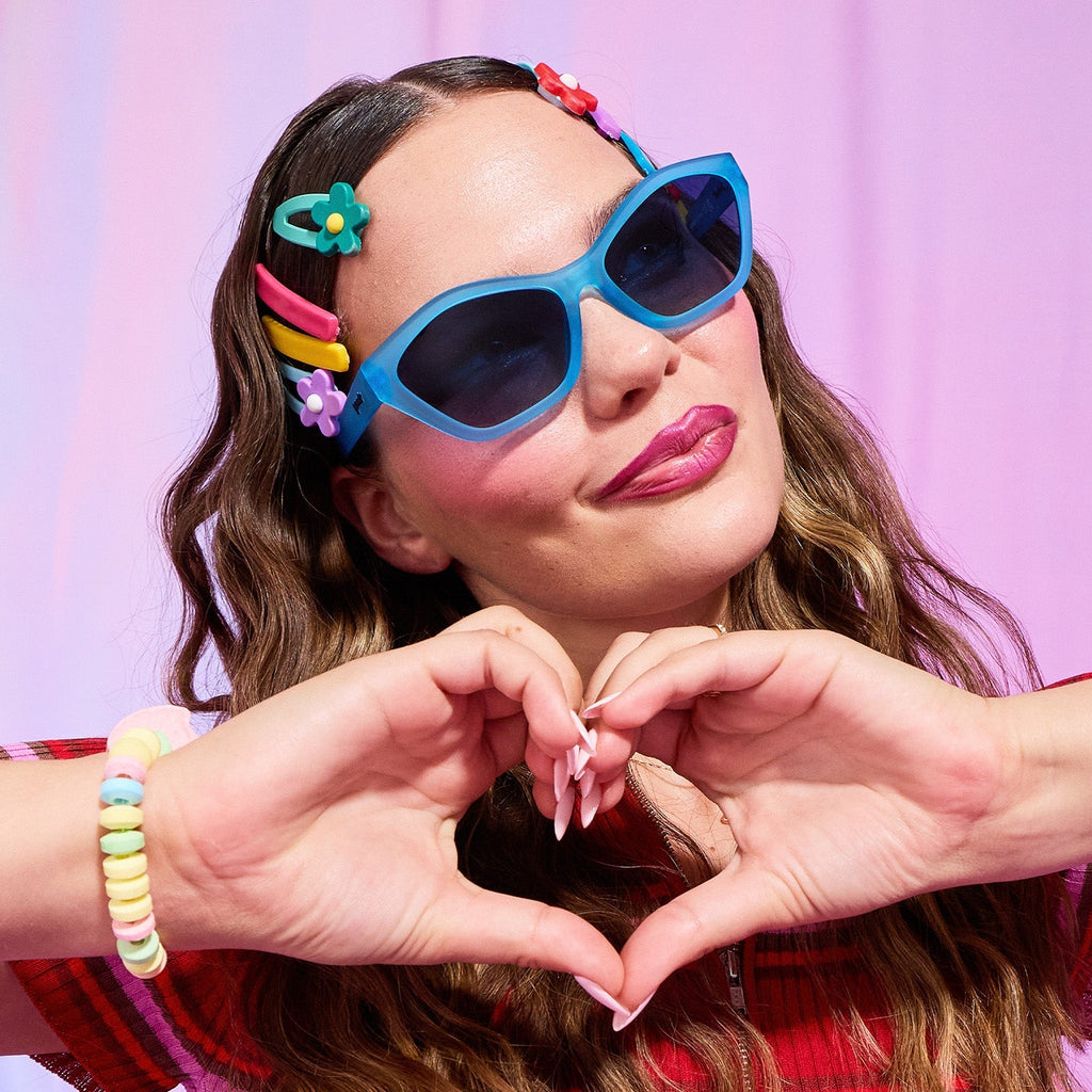 A young brunette woman wearing blue Deco G sunglasses, candy bracelets, and colorful barettes makes a heart with her hands.