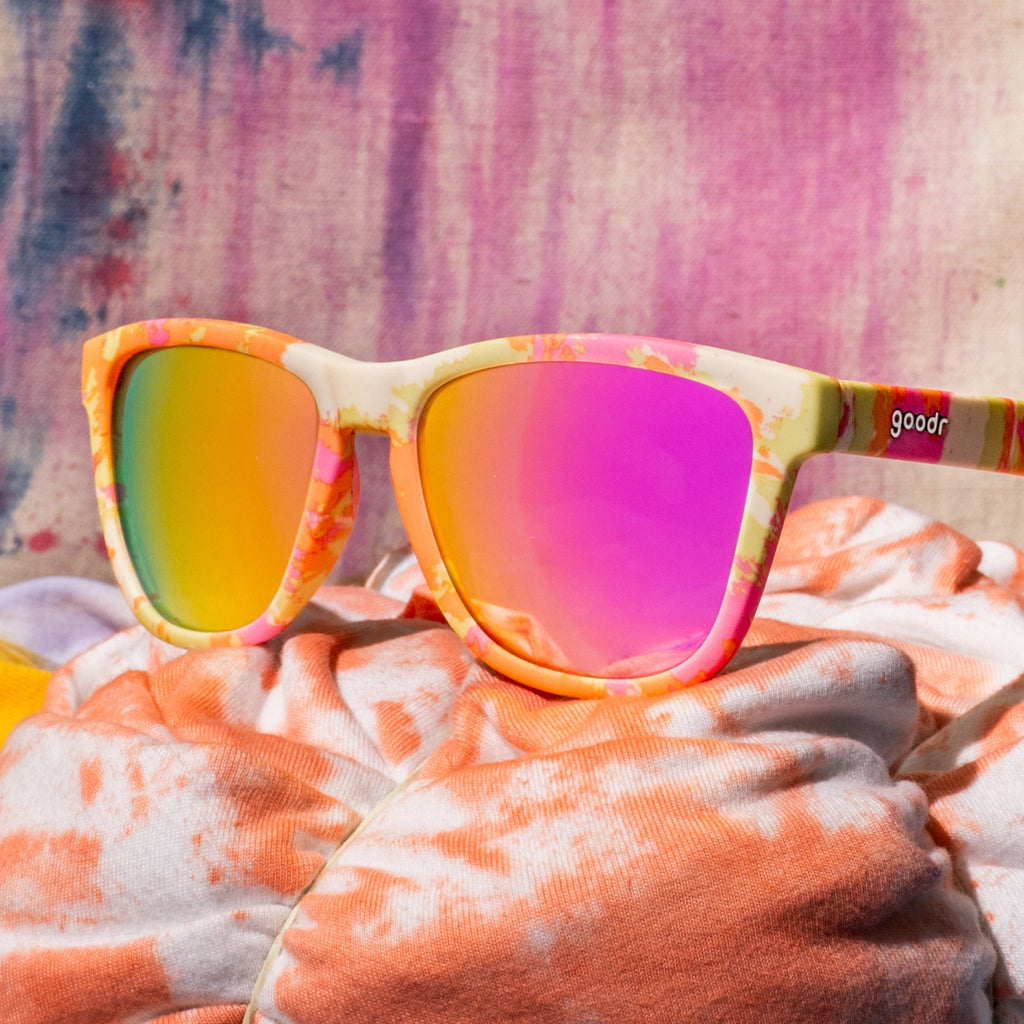 Yellow, pink, and orange tie-dye shades with pink reflective lenses sitting on white and orange tie-dyed fabric.