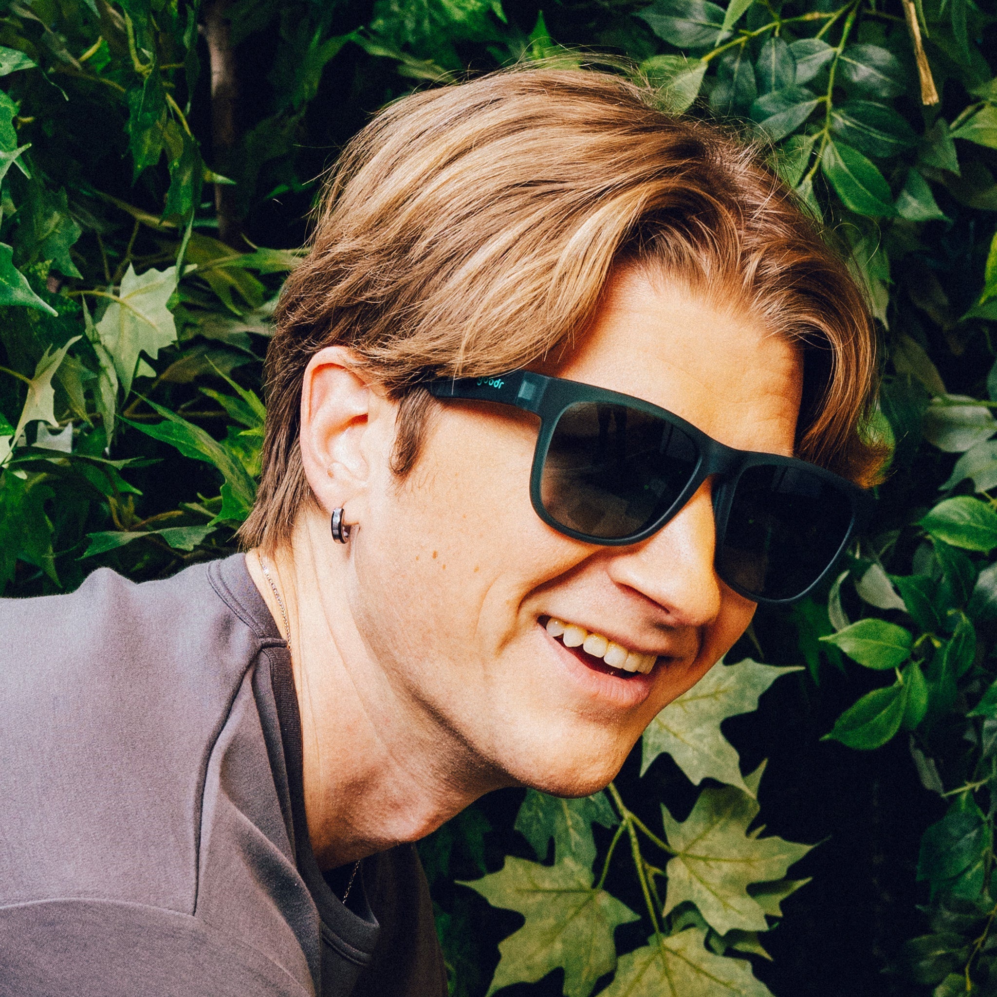 Man poses in front of vines, wearing large dark teal sunglasses with green gradient lenses.