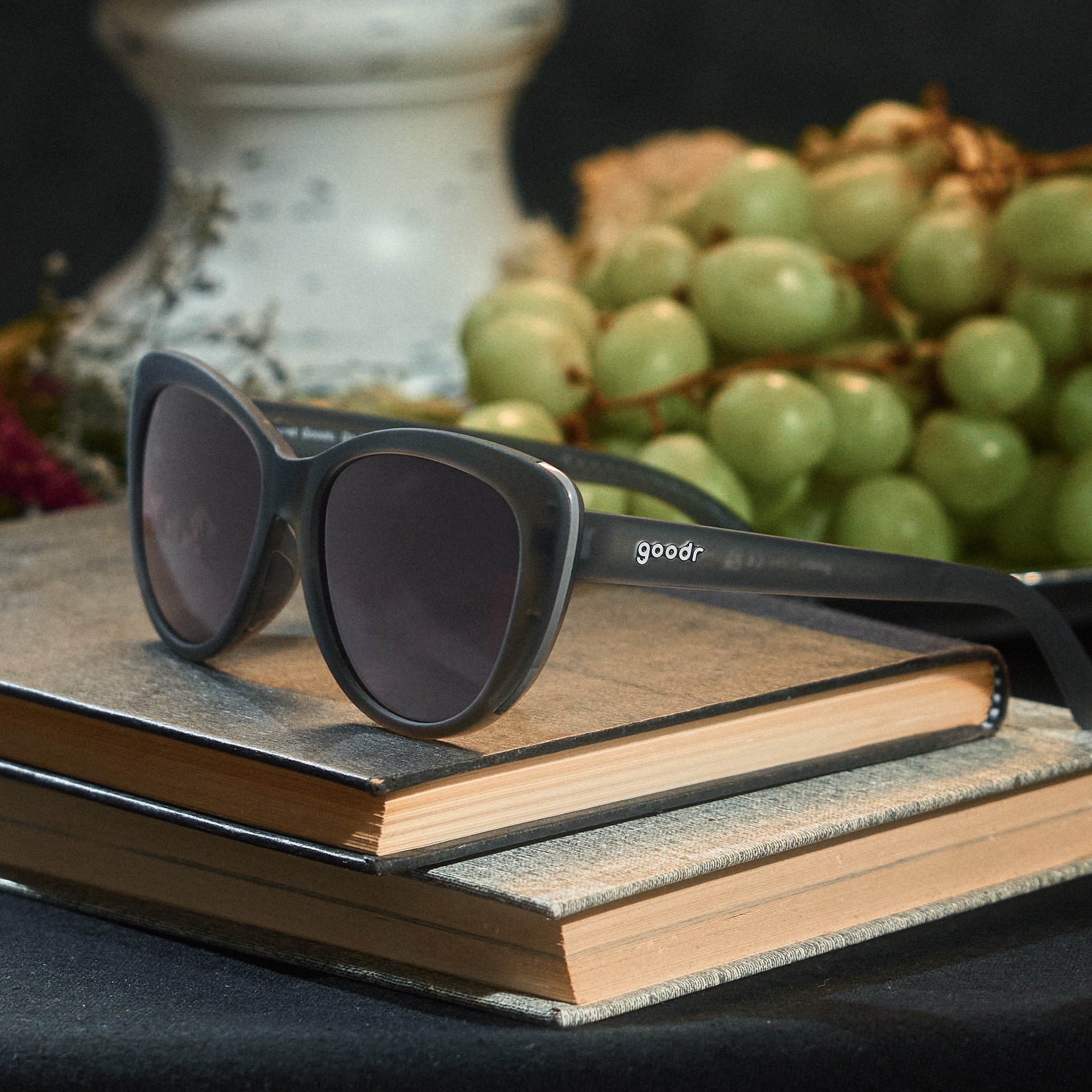 Three-quarter angle view of translucent gray cat-eye sunglasses with black gradient lenses on top of stacked books.