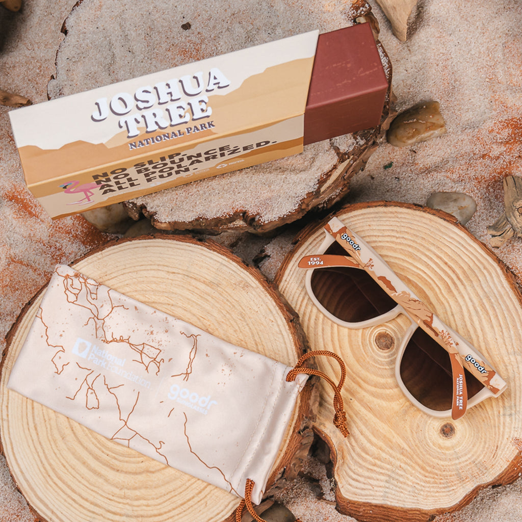 Joshua Tree National Park-themed sunglasses with brown-tinted lenses and a desert landscape design on the frames, displayed on a wood slice with a branded carrying pouch and box.