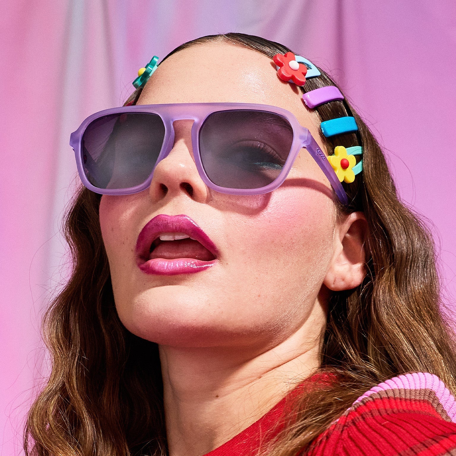A young brunette woman with multiple candy-colored barettes models translucent purple Retro G shades against a pink backdrop.