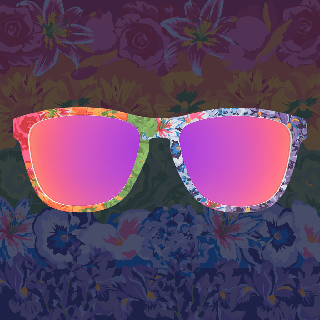 Front view of colorful floral print goodr sunglasses with purple reflective sunglasses against a floral background.