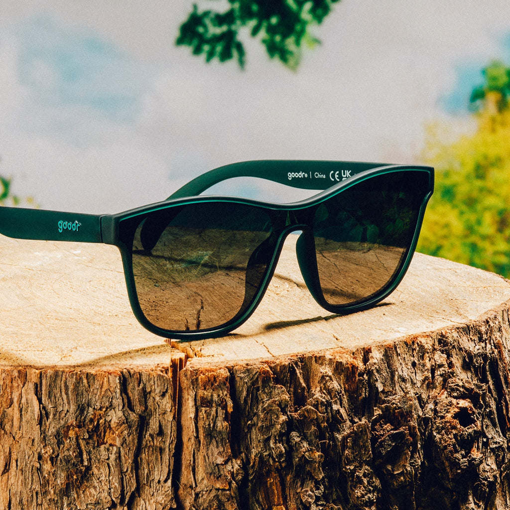 Three-quarter angle view of translucent teal one-lens sunglasses with green gradient lenses on a tree trunk in front of a nature backdrop.