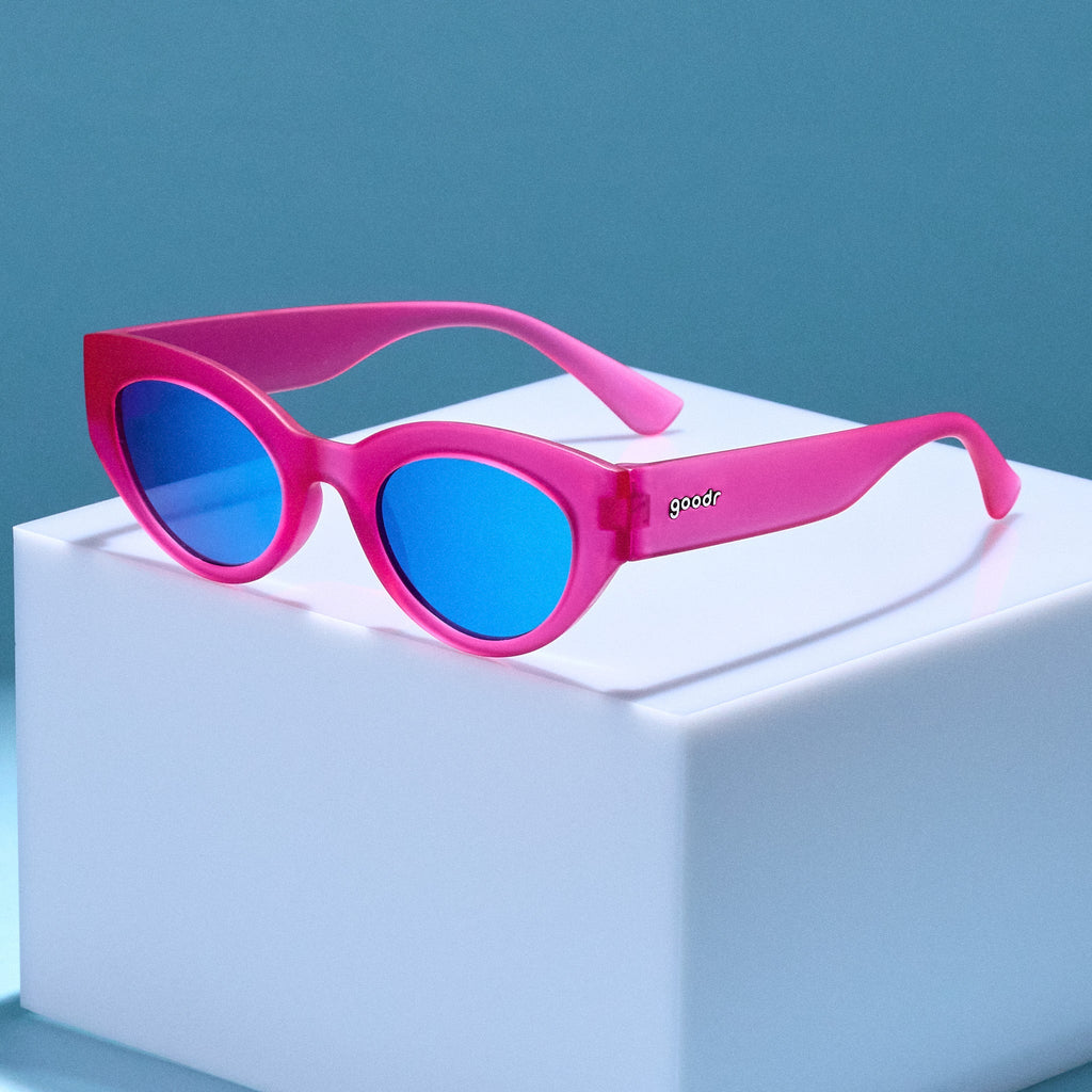 A pair of translucent hot pink cat-eye frames with blue reflective lenses posed on a white cube against a blue backdrop.