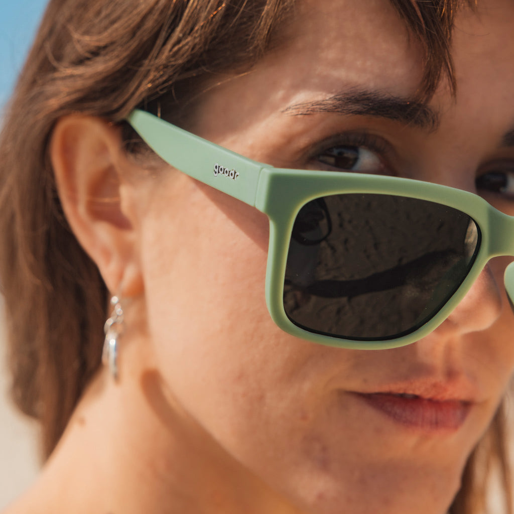 Three-quarter angle view of a composed young woman with dark hair wearing green sunglasses designed to float in water.