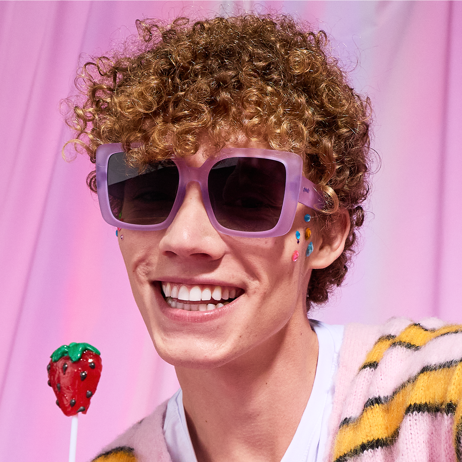 A young man with curly, strawberry-blonde hair smiles enthusiastically and models oversized, pale purple square sunnies.