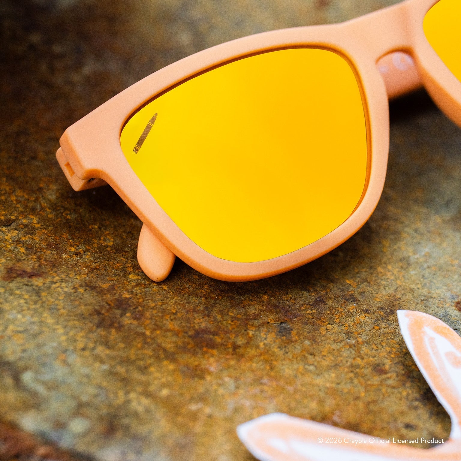Close-up angled view of tan sunglasses w/ amber reflective lenses + small crayon etching in top corner of lens.