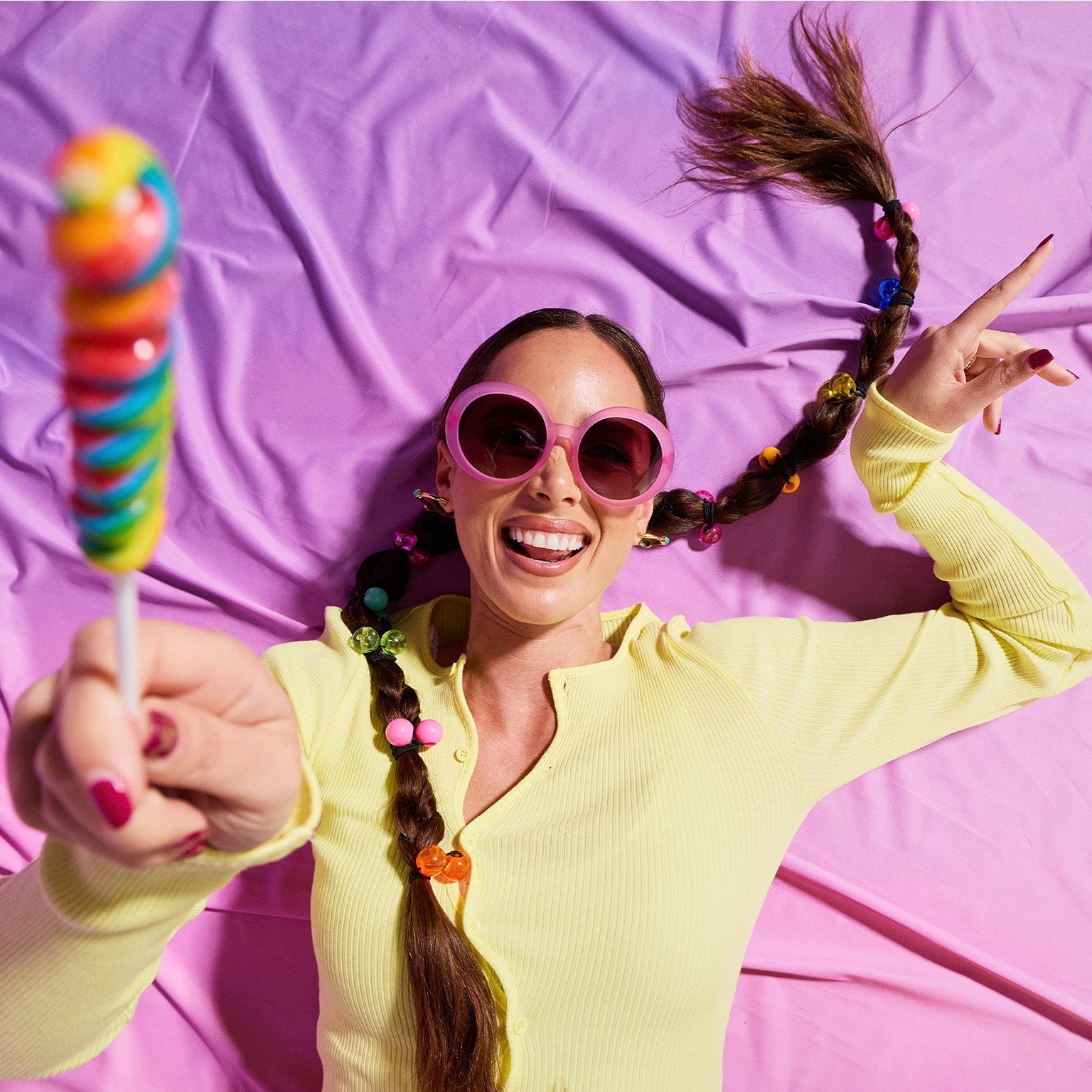A dark-pigtailed woman lies on a purplish-pink backdrop in translucent-pink Mood G sunnies, holding a rainbow twirl lollipop.