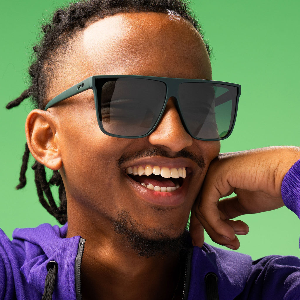 Closeup of a stylish man smiling gleefully and rocking dark green oversized flat-top sunglasses with green gradient lenses.
