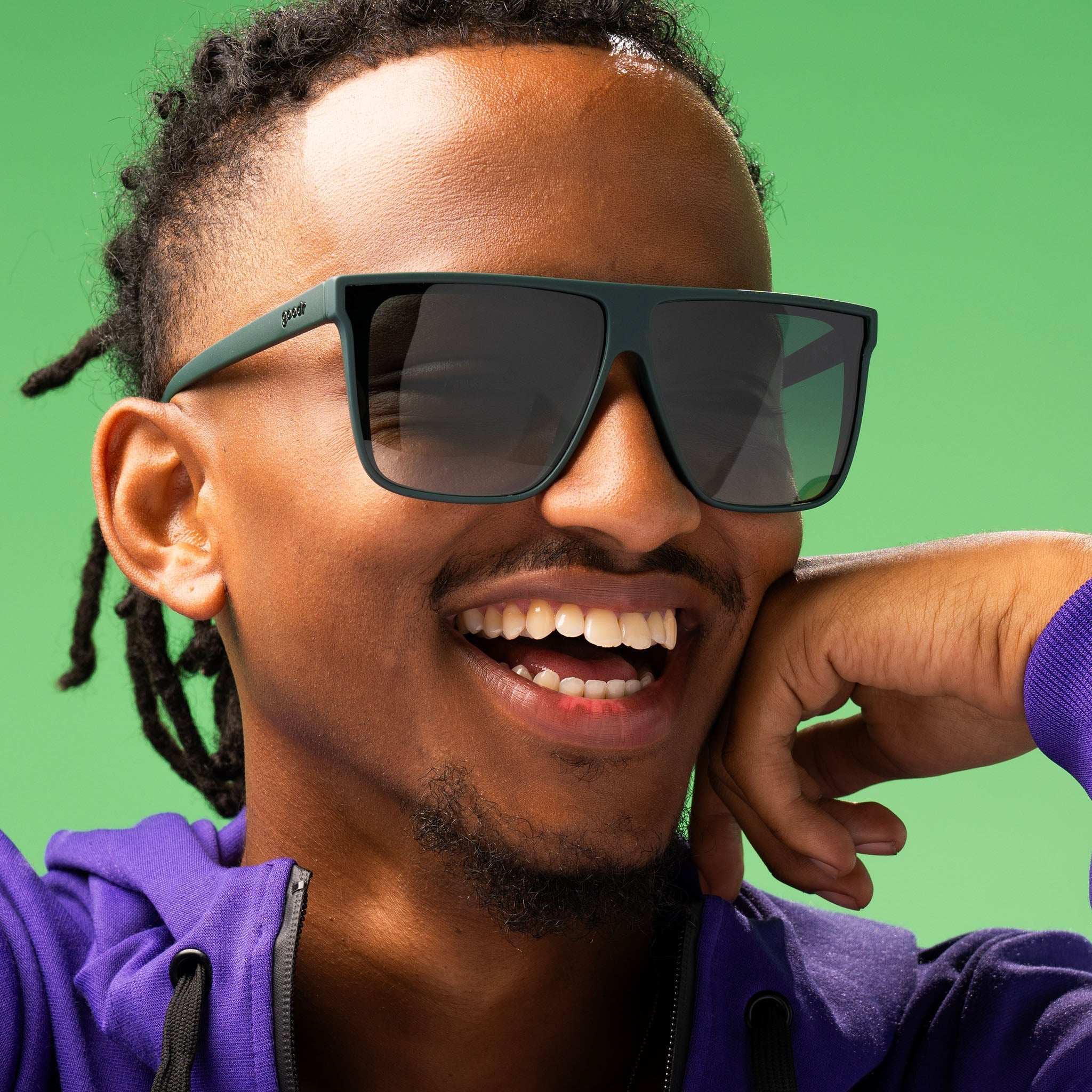 Closeup of a stylish man smiling gleefully and rocking dark green oversized flat-top sunglasses with green gradient lenses.