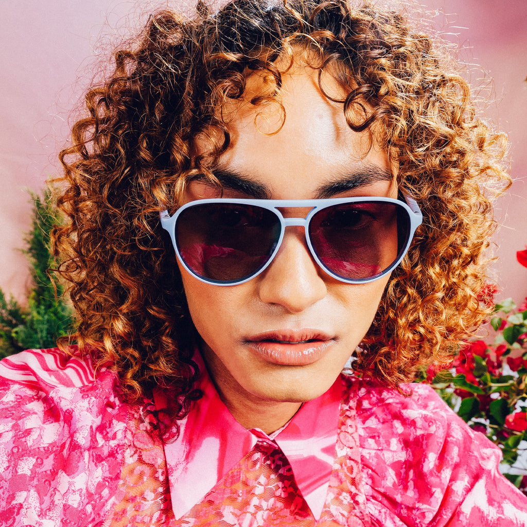 Front view of woman wearing light blue aviator sunglasses with purple gradient lenses against a meadow backdrop.