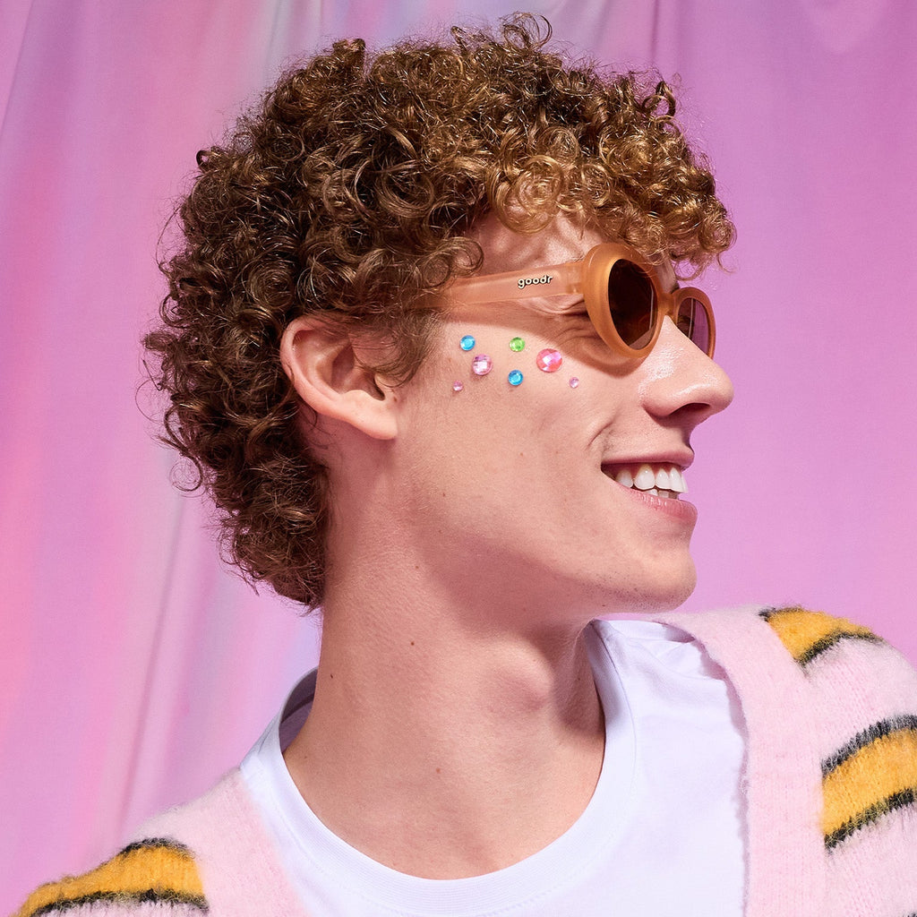 Profile of a young man with strawberry blond curls and rhinestones on his cheek smiles in a pair of peach Beat G shades.