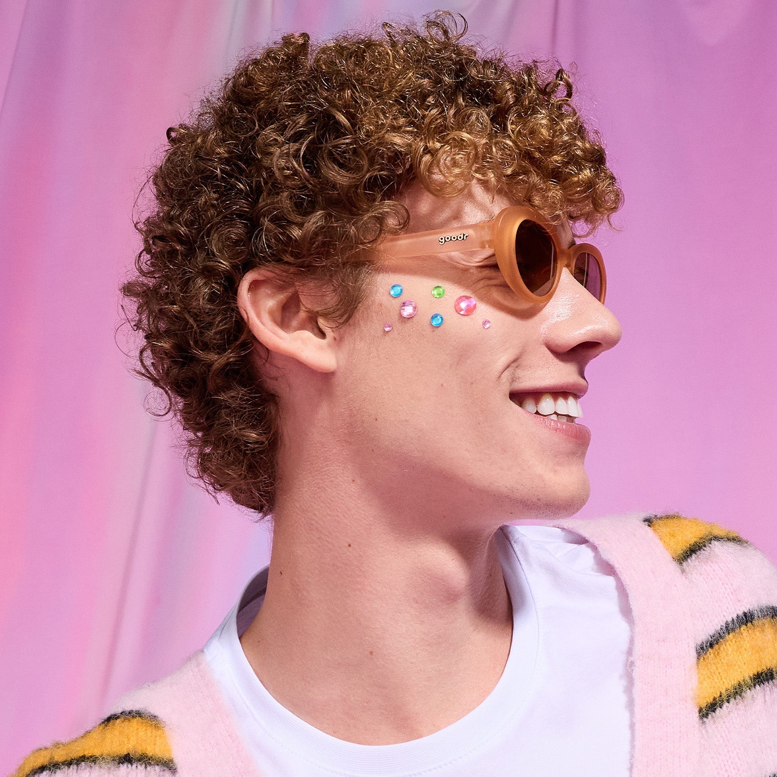 Profile of a young man with strawberry blond curls and rhinestones on his cheek smiles in a pair of peach Beat G shades.