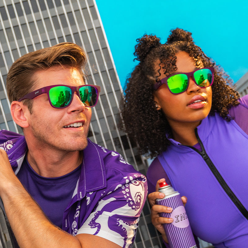 A man and a woman wearing purple clothing and reflective sunglasses with green lenses and purple frames.