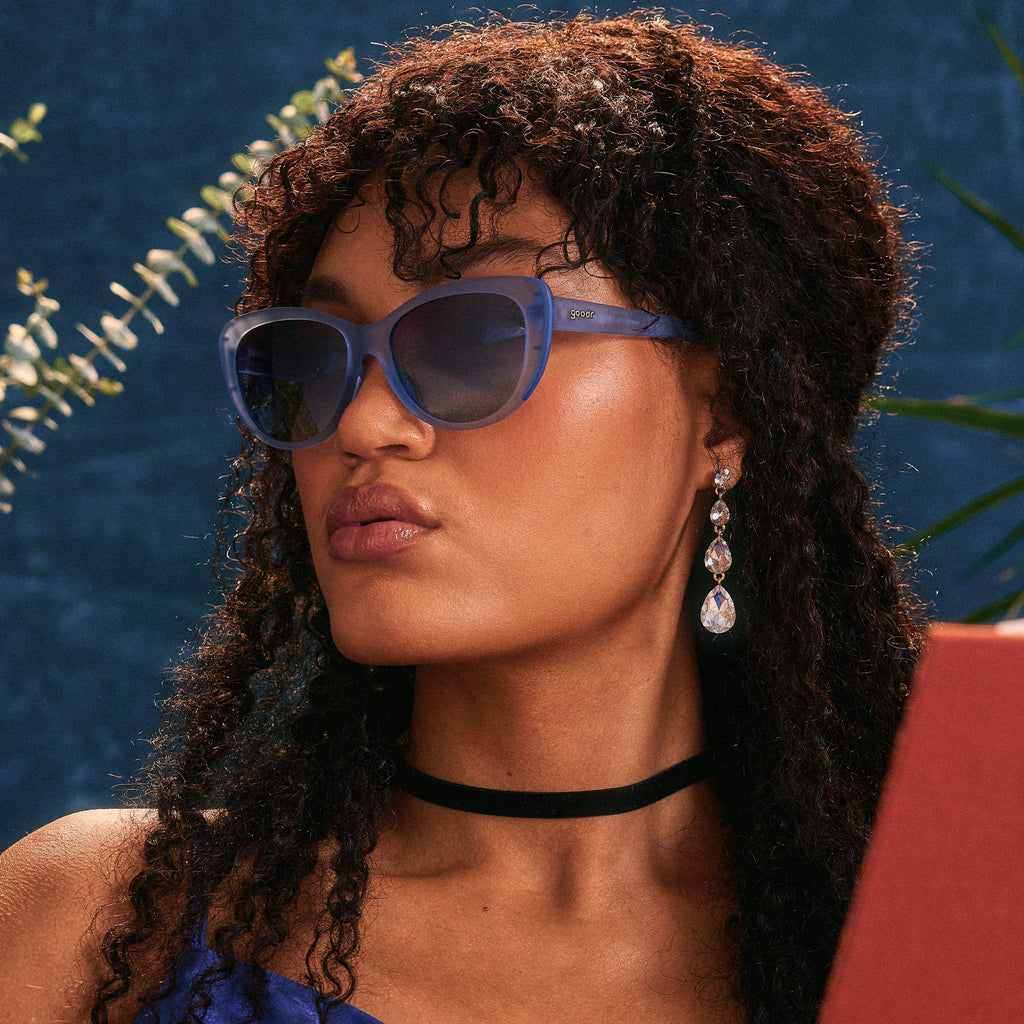 A young woman with long, dark, curly hair poses in a pair of dusty blue translucent Glam Gs against a cerulean background.