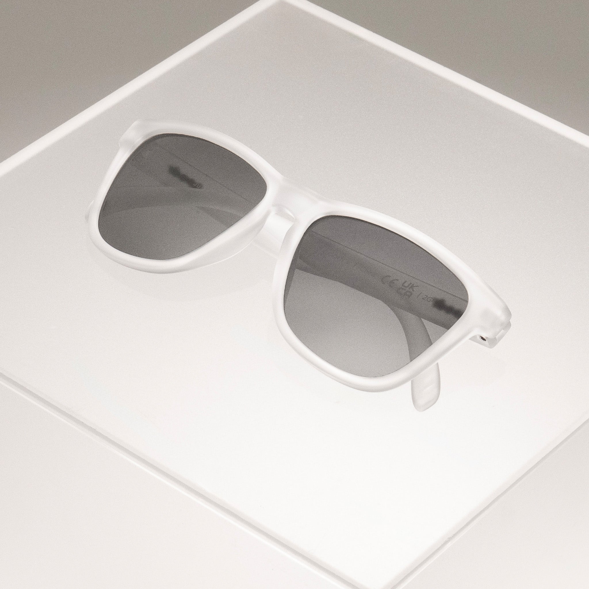 Clear sunglasses in a classic frame shape with black non-reflective lenses folded, sitting atop a clear plastic box.