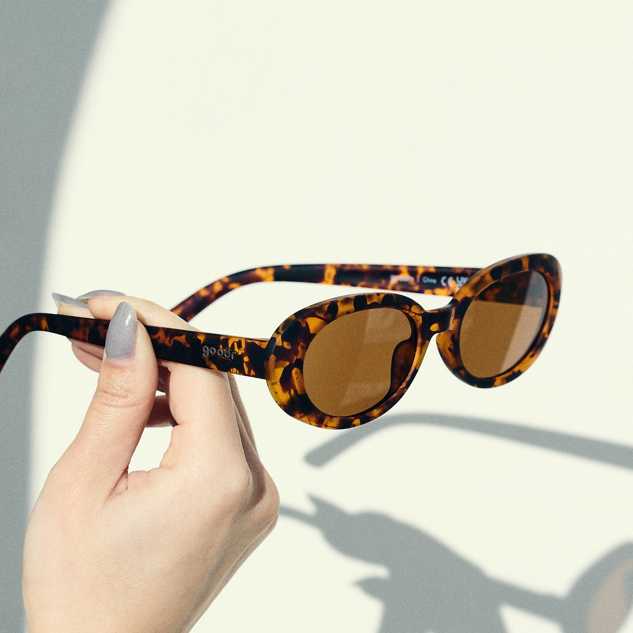 A manicured hand holds a pair of oval tortoiseshell sunglasses with brown polarized lenses in front of an off-white backdrop.