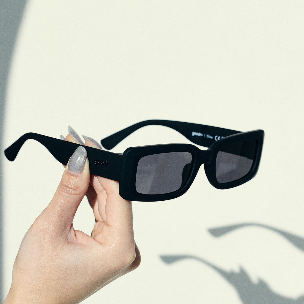 A manicured hand holds up a pair of black rectangular sunglasses with black polarized lenses against an off-white backdrop.