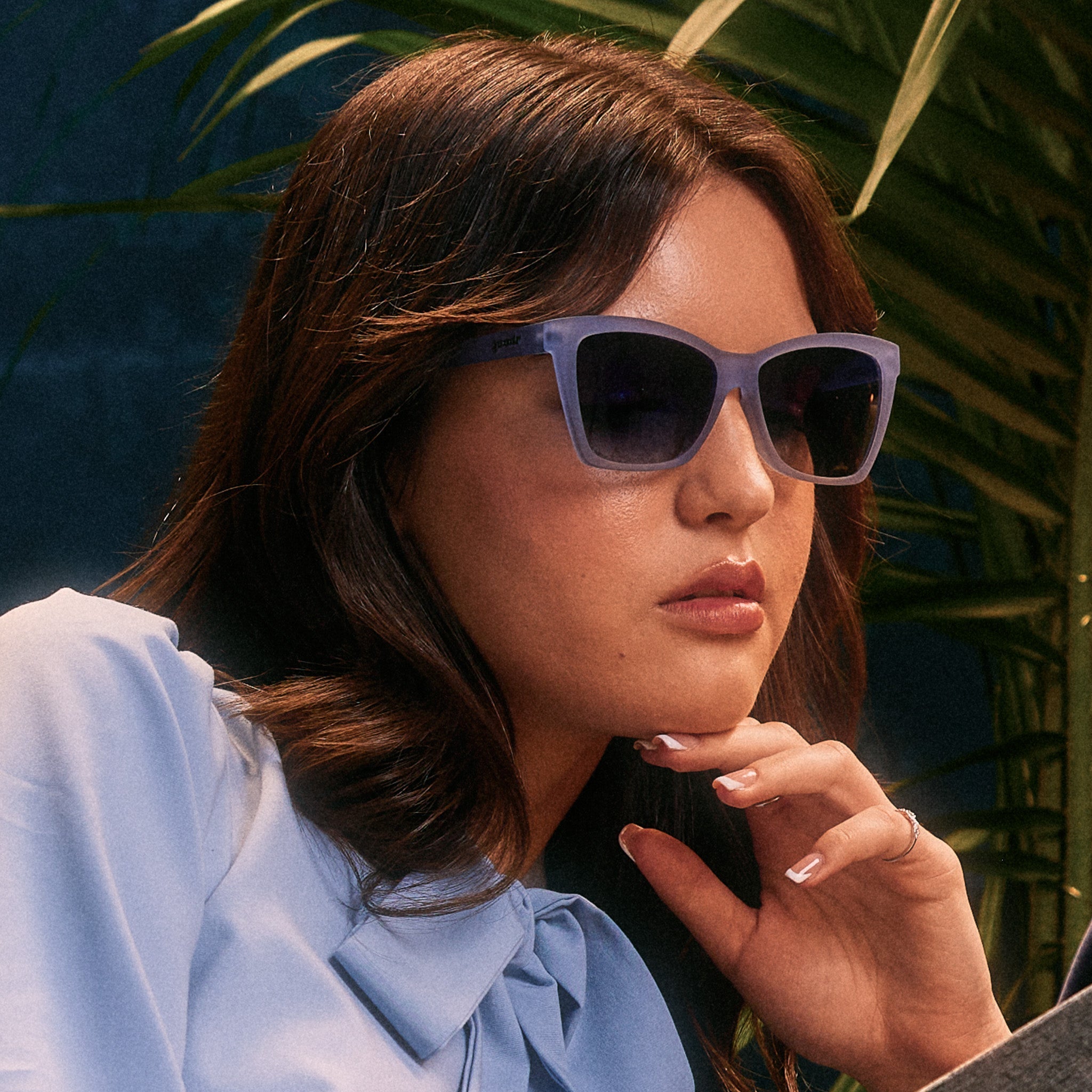 A woman in a pale blue pussybow blouse wears translucent blue Pop Gs and is resting her chin thoughtfully on her hand.
